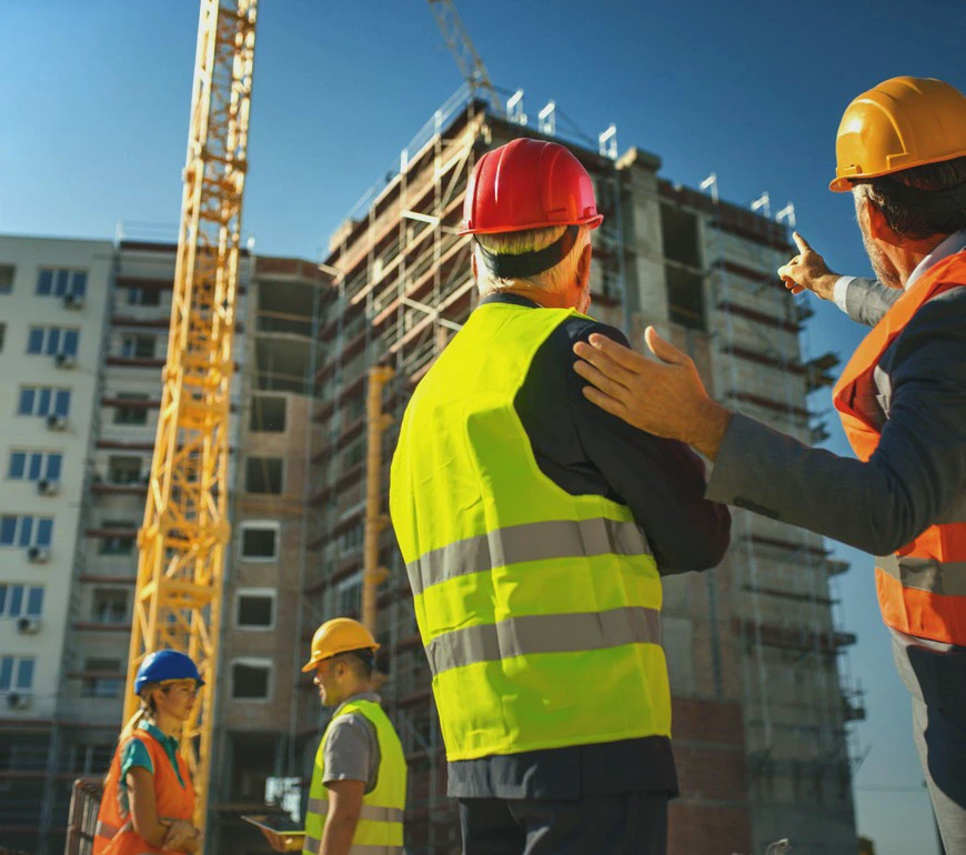 Pracownicy budowlani w&nbsp;kaskach i&nbsp;kamizelkach odblaskowych na&nbsp;placu budowy, na&nbsp;tle rusztowań i&nbsp;dźwigu