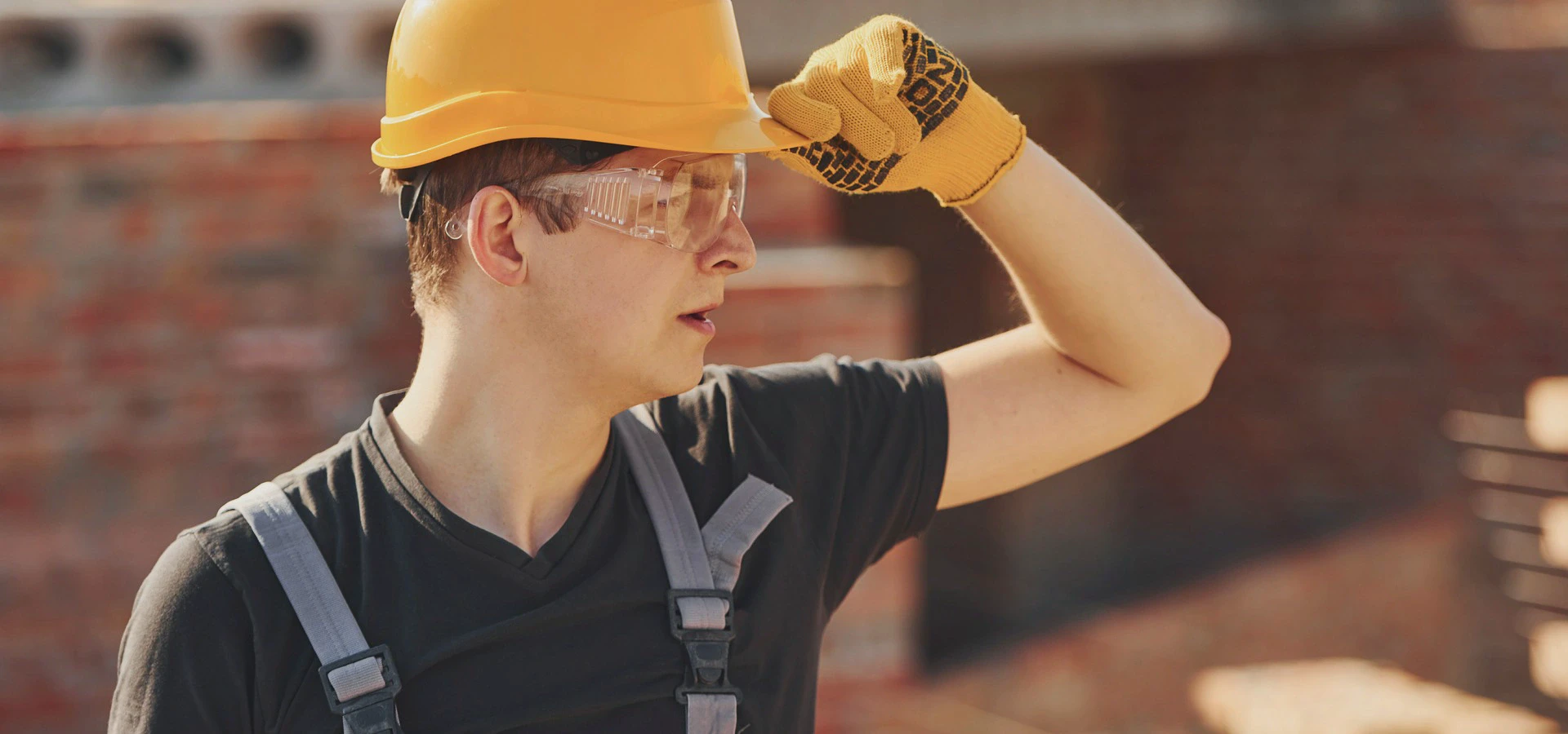 Osoba w&nbsp;kasku ochronnym i&nbsp;rękawicach roboczych stoi na&nbsp;placu budowy, osłaniając dłonią oczy przed światłem
